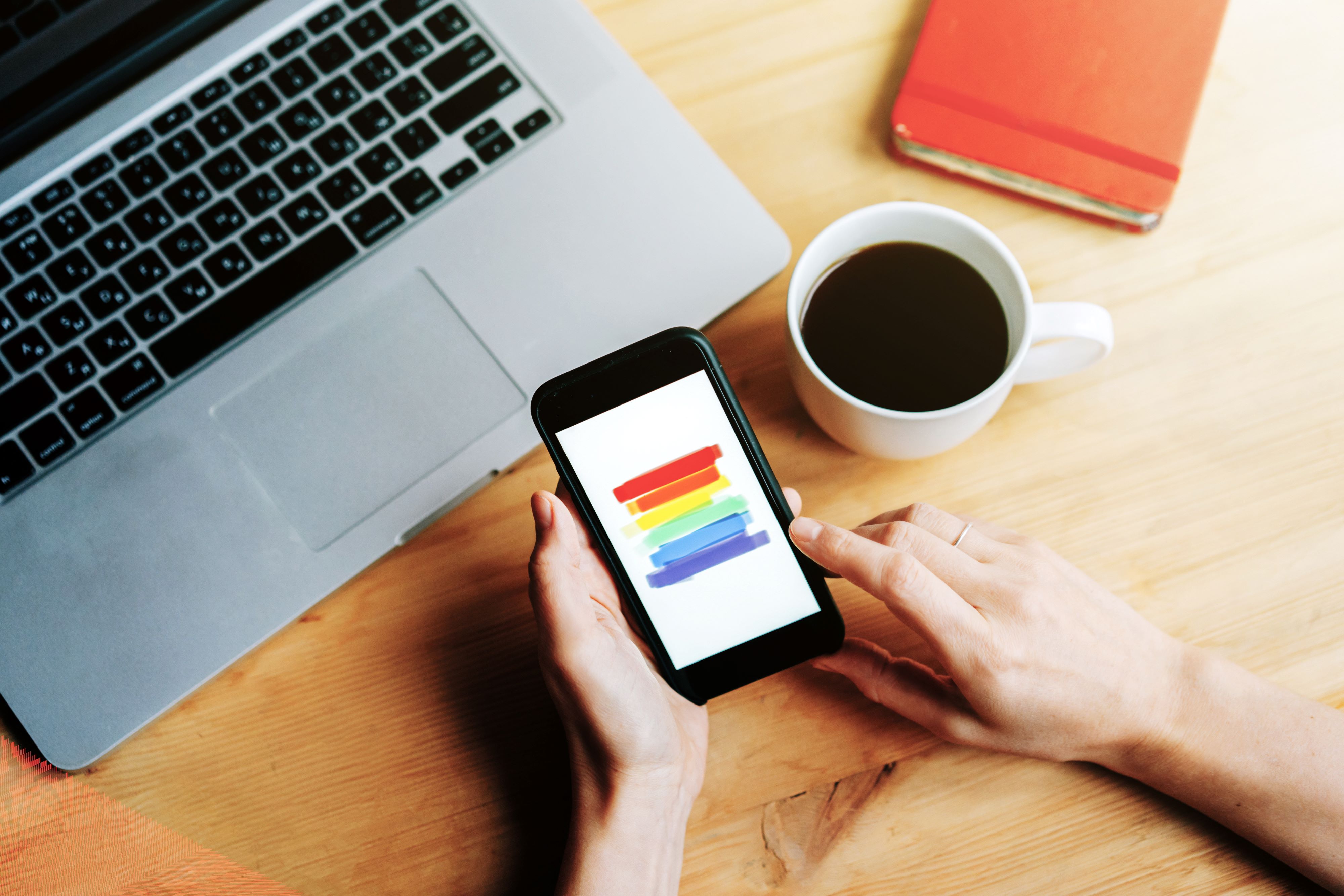 Person using phone in office, laptop and coffee, with LGBT+ flag on screen